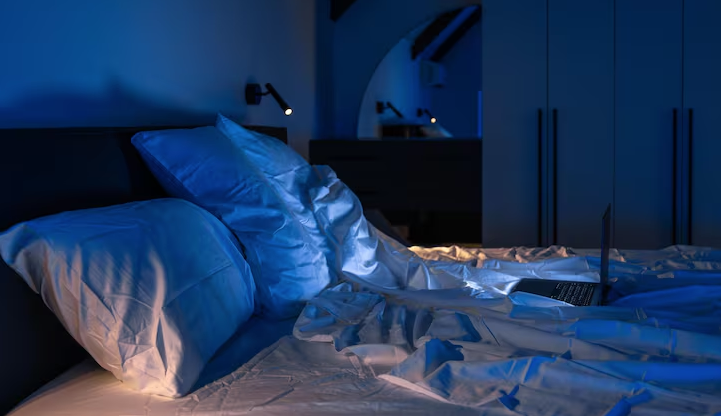 Unmade bed with an open laptop in a blue-lit bedroom at night